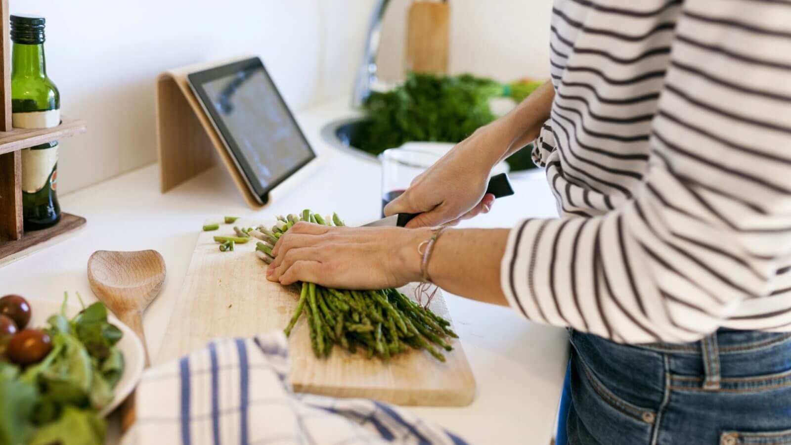 Frau schneidet grünen Spargel auf einem Brett. Daneben steht ein Tablet.